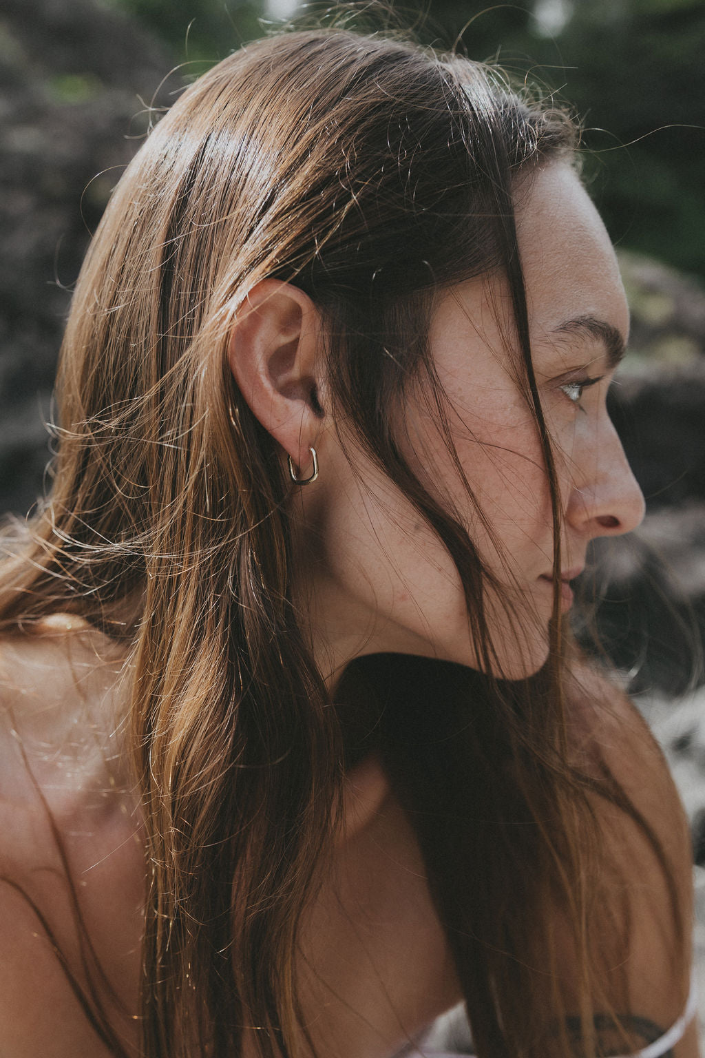 A close-up side profile of a woman with long brown hair wearing a small gold hoop earring, set against a blurred natural outdoor background.