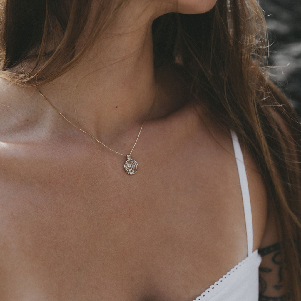 A close-up lifestyle shot of a woman wearing a delicate gold chain necklace with a small circular pendant, paired with a white spaghetti strap top.