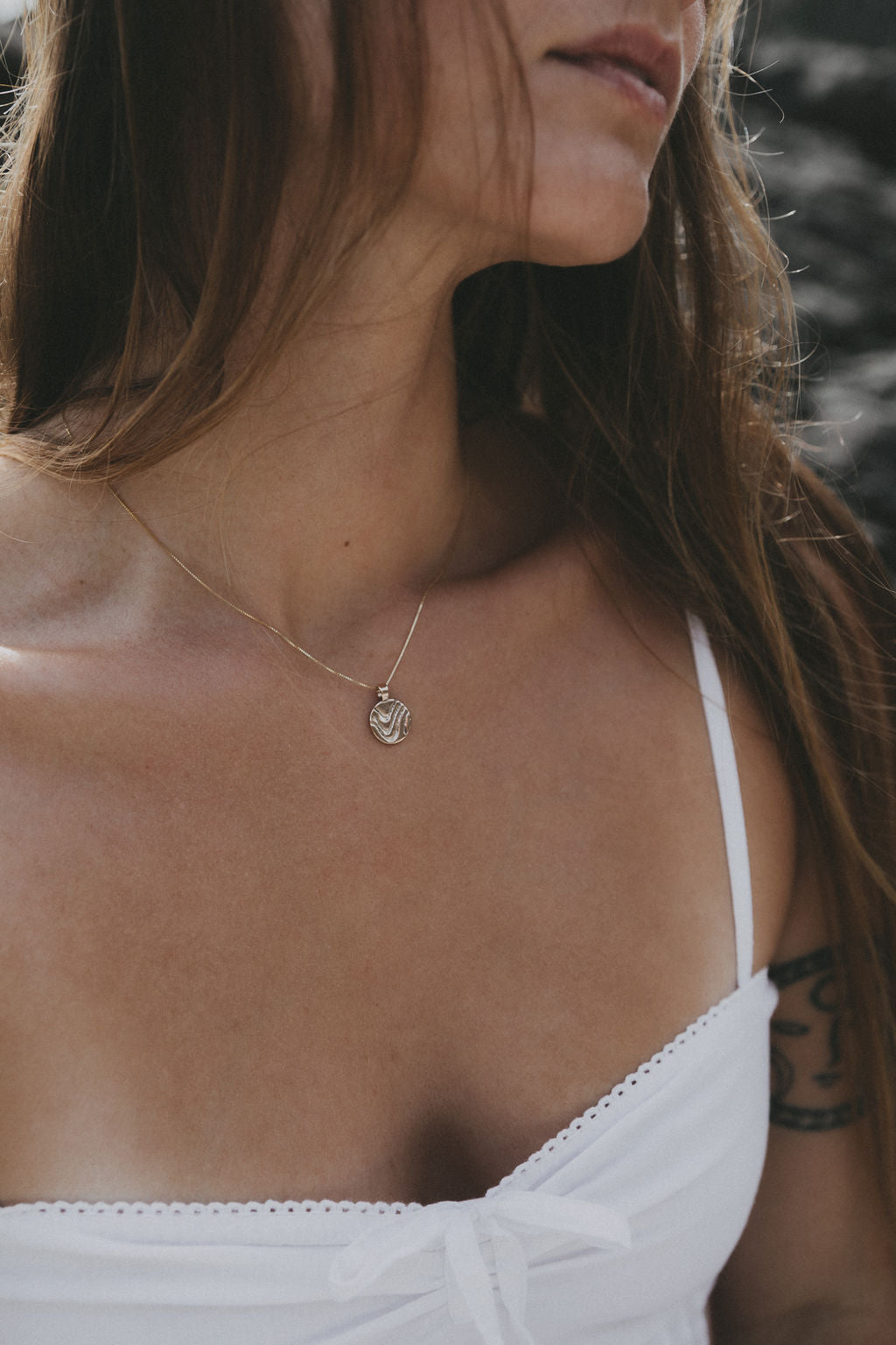 A close-up lifestyle shot of a woman wearing a delicate gold chain necklace with a small circular pendant, paired with a white spaghetti strap top.