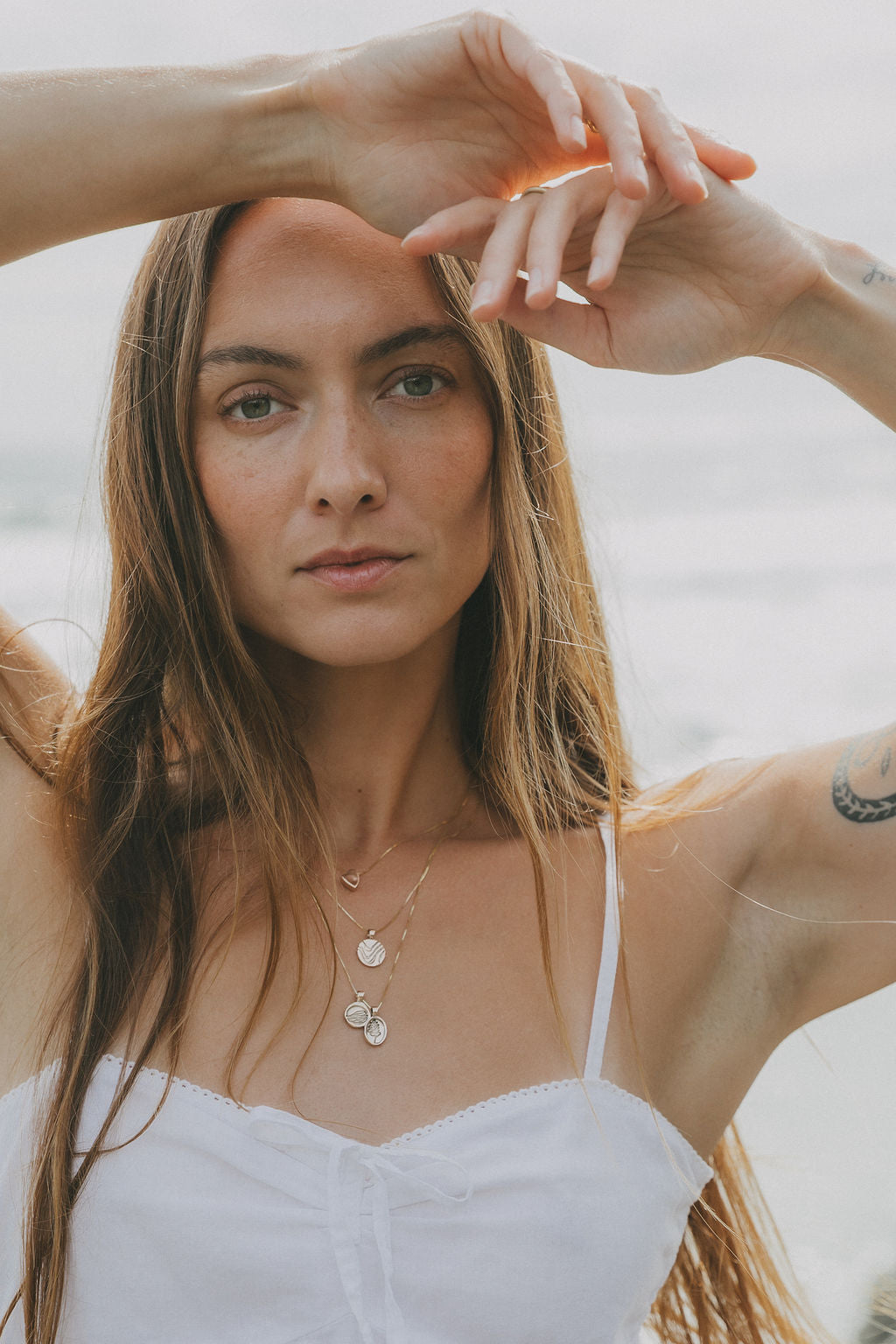 A woman with long light brown hair wearing a white spaghetti strap top and layered gold necklaces, posing with her hands near her face against a soft, blurred beach background.