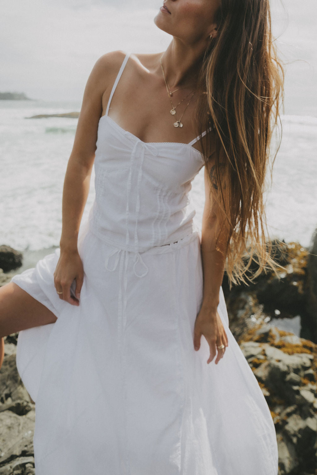 A woman wearing a white spaghetti-strap maxi dress stands on a rocky beach with the ocean in the background.
