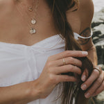 A close-up lifestyle shot of a woman wearing layered gold and rose gold necklaces, a gold ring, and a face mask, while holding her brown hair outdoors near water.