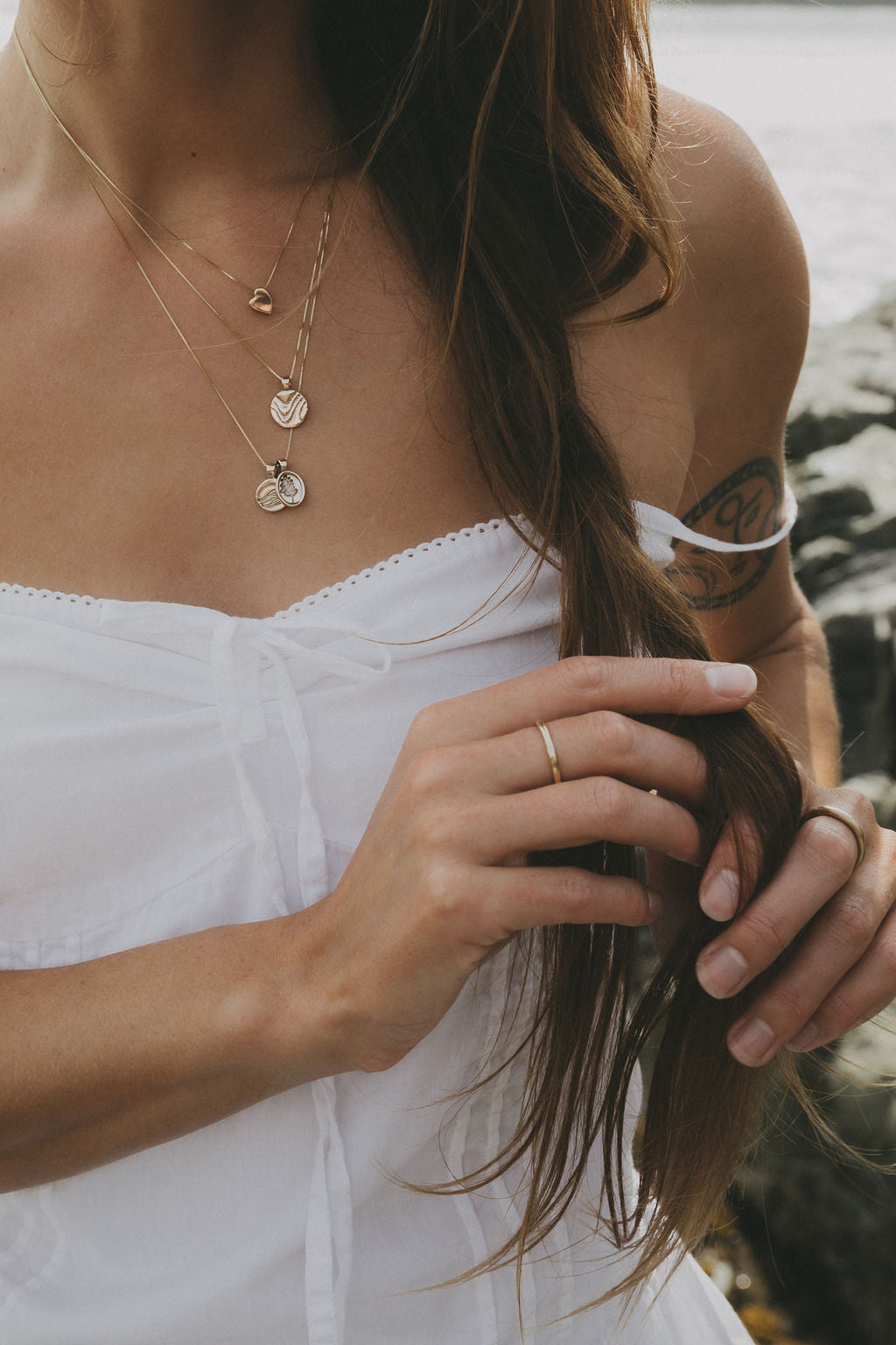 A close-up lifestyle shot of a woman wearing layered gold and rose gold necklaces, a gold ring, and a face mask, while holding her brown hair outdoors near water.