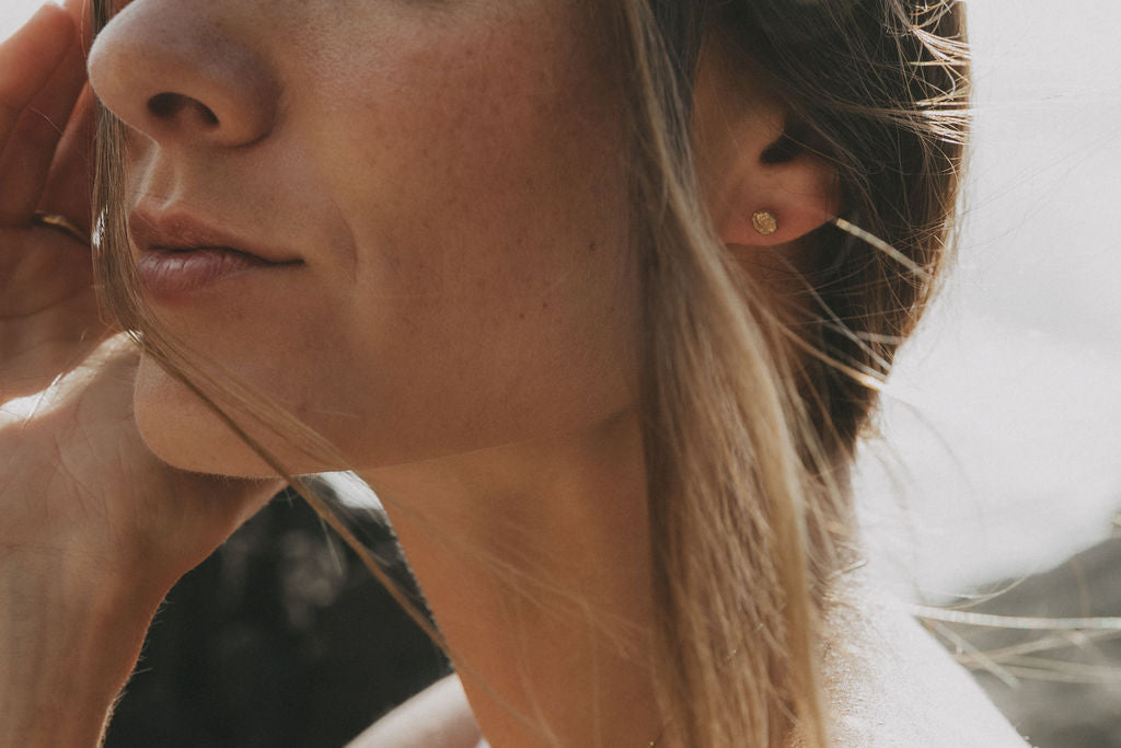 Close-up of a woman's ear wearing a small gold stud earring, with her face partially visible and soft natural light on her skin and hair.