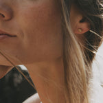 Close-up of a woman's ear wearing a small gold stud earring, with her face partially visible and soft natural light on her skin and hair.