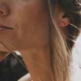 Close-up of a woman's ear wearing a small gold stud earring, with her face partially visible and soft natural light on her skin and hair.