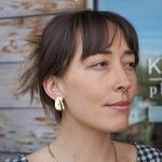 Woman with a nose ring and gold earring and necklace, wearing a checkered top, standing outdoors.