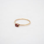 A delicate yellow gold ring featuring a small, round-cut dark red gemstone in a prong setting, photographed on a clean white background.