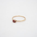 A delicate yellow gold ring featuring a small, round-cut dark red gemstone in a prong setting, photographed on a clean white background.