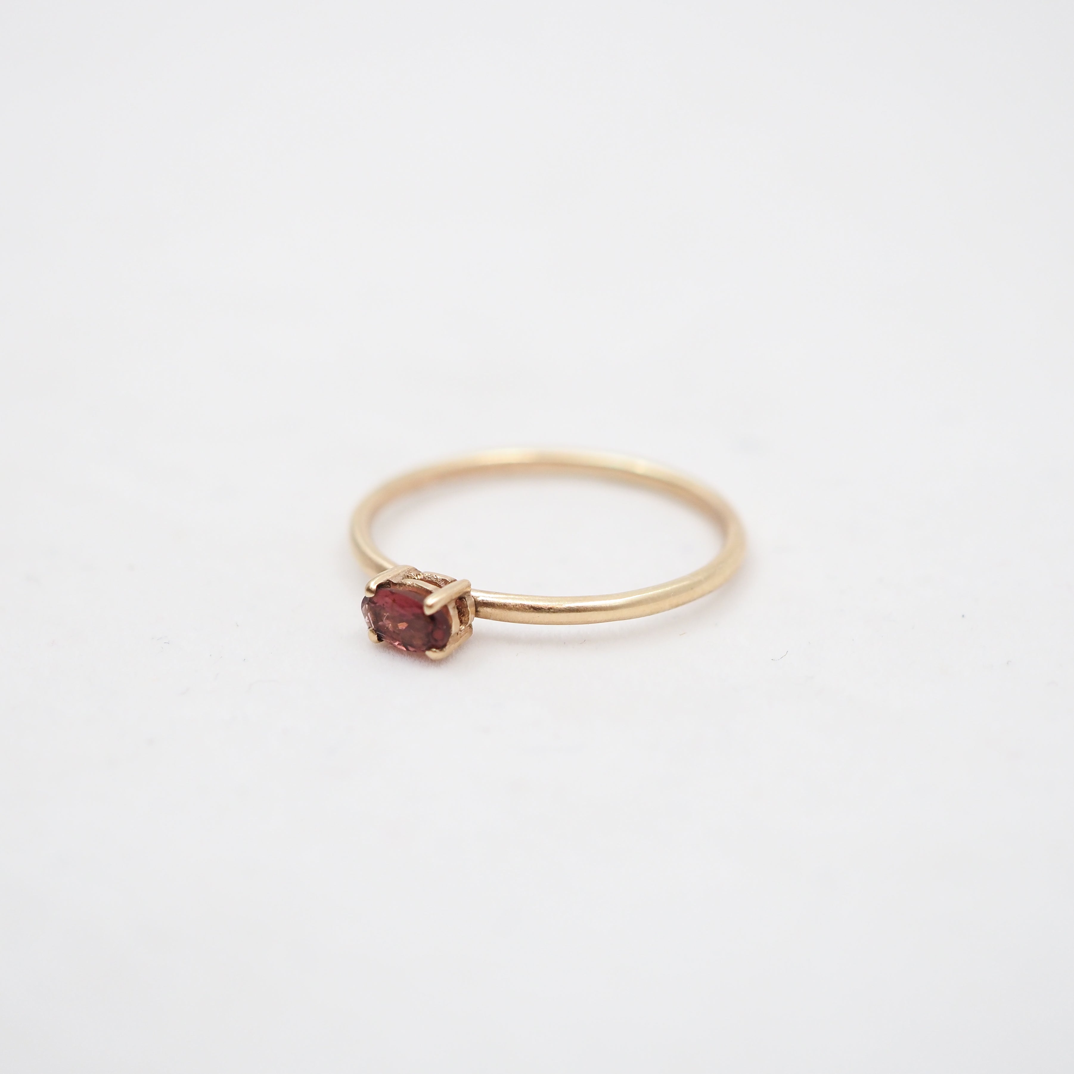 A delicate yellow gold ring featuring a small, round-cut dark red gemstone in a prong setting, photographed on a clean white background.