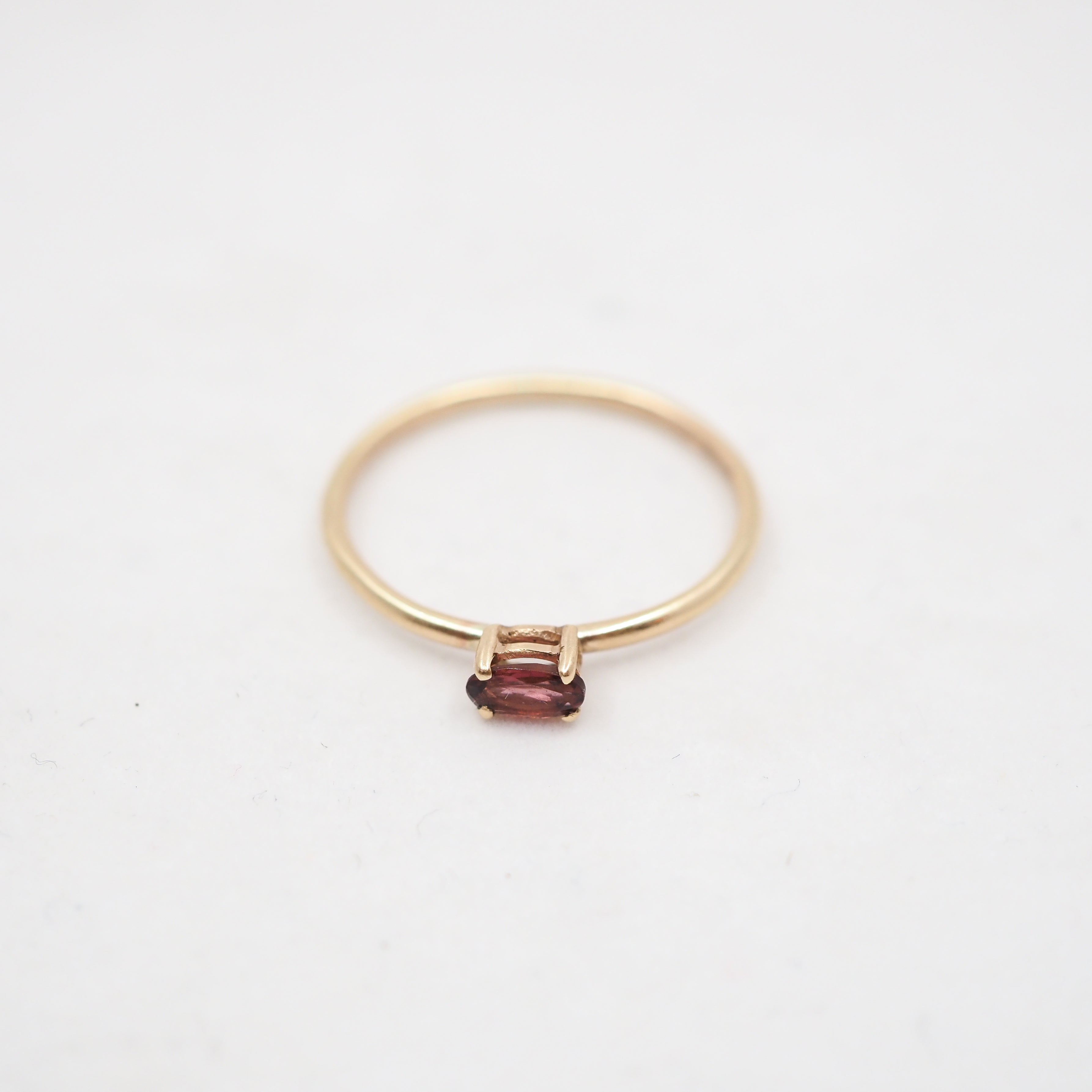 A delicate yellow gold ring featuring a small, rectangular-cut dark red gemstone in a four-prong setting, photographed on a clean white background.