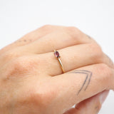 A close-up of a hand wearing a delicate gold ring with a small oval pink gemstone, set against a plain white background.