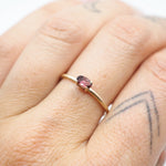 A close-up of a hand wearing a delicate gold ring with a small oval pink gemstone.