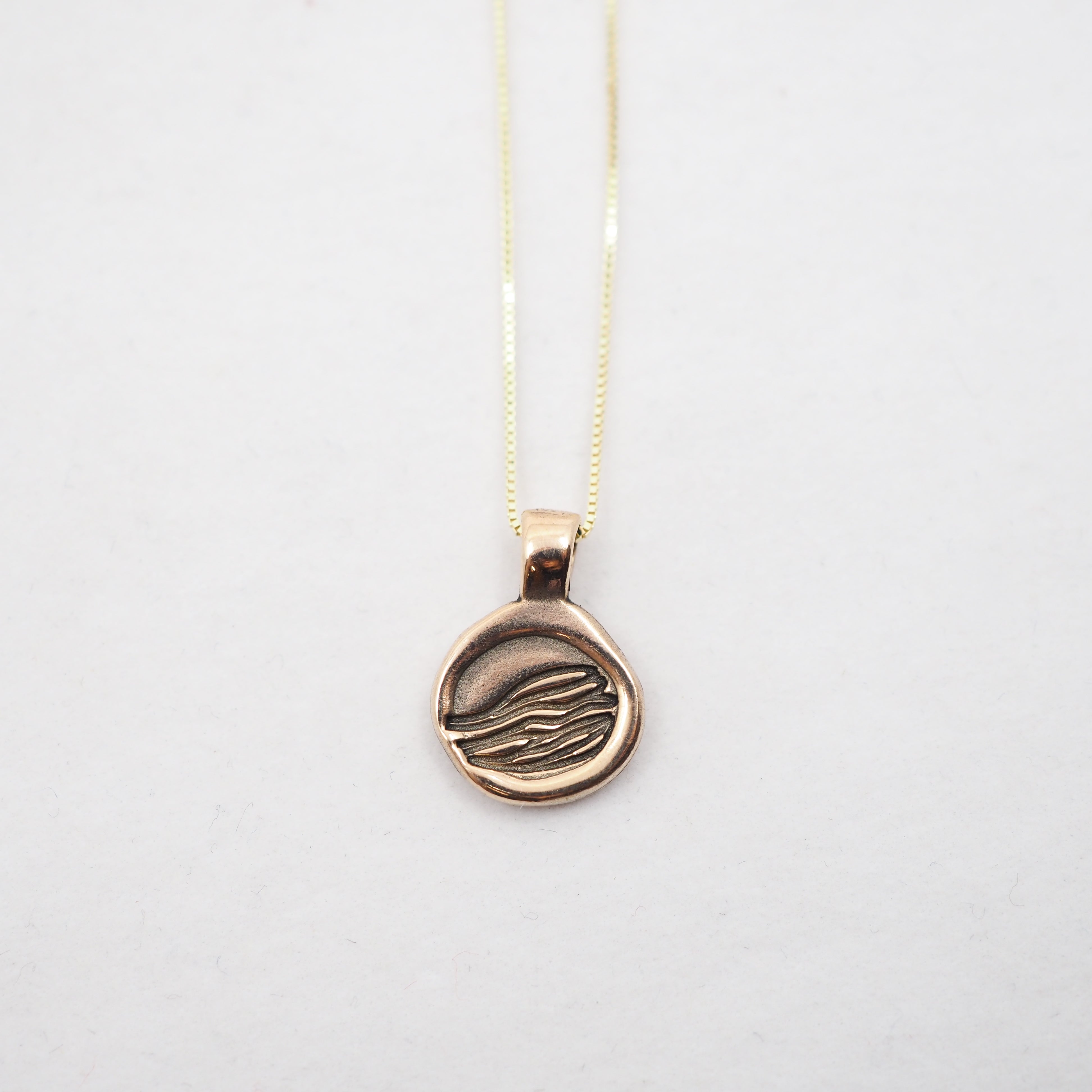 A gold pendant necklace featuring a circular disc pendant with an engraved stylized hand design, suspended from a delicate gold chain against a plain white background.