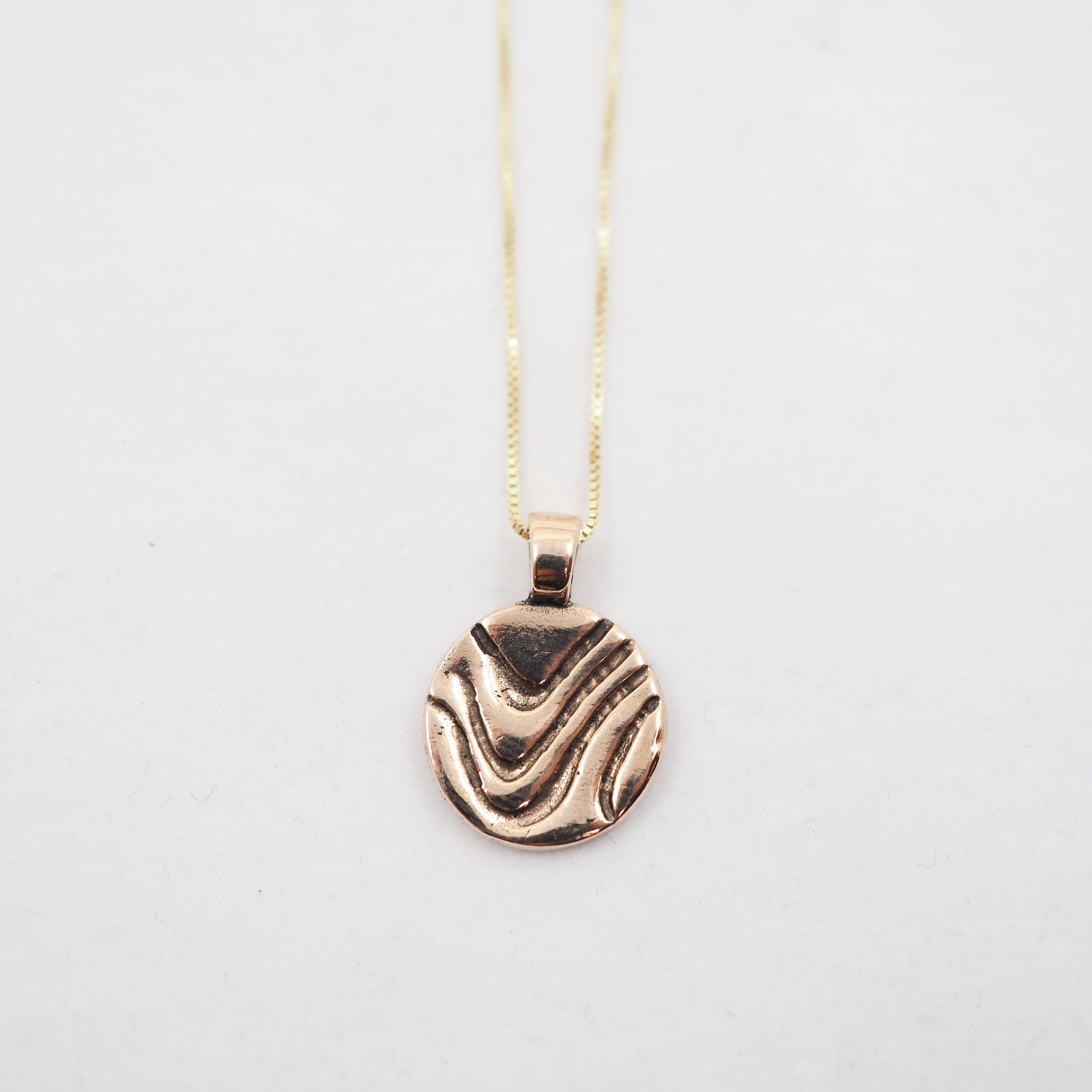 A gold pendant necklace featuring a textured, wavy circular disc pendant, suspended from a delicate gold chain against a plain white background.