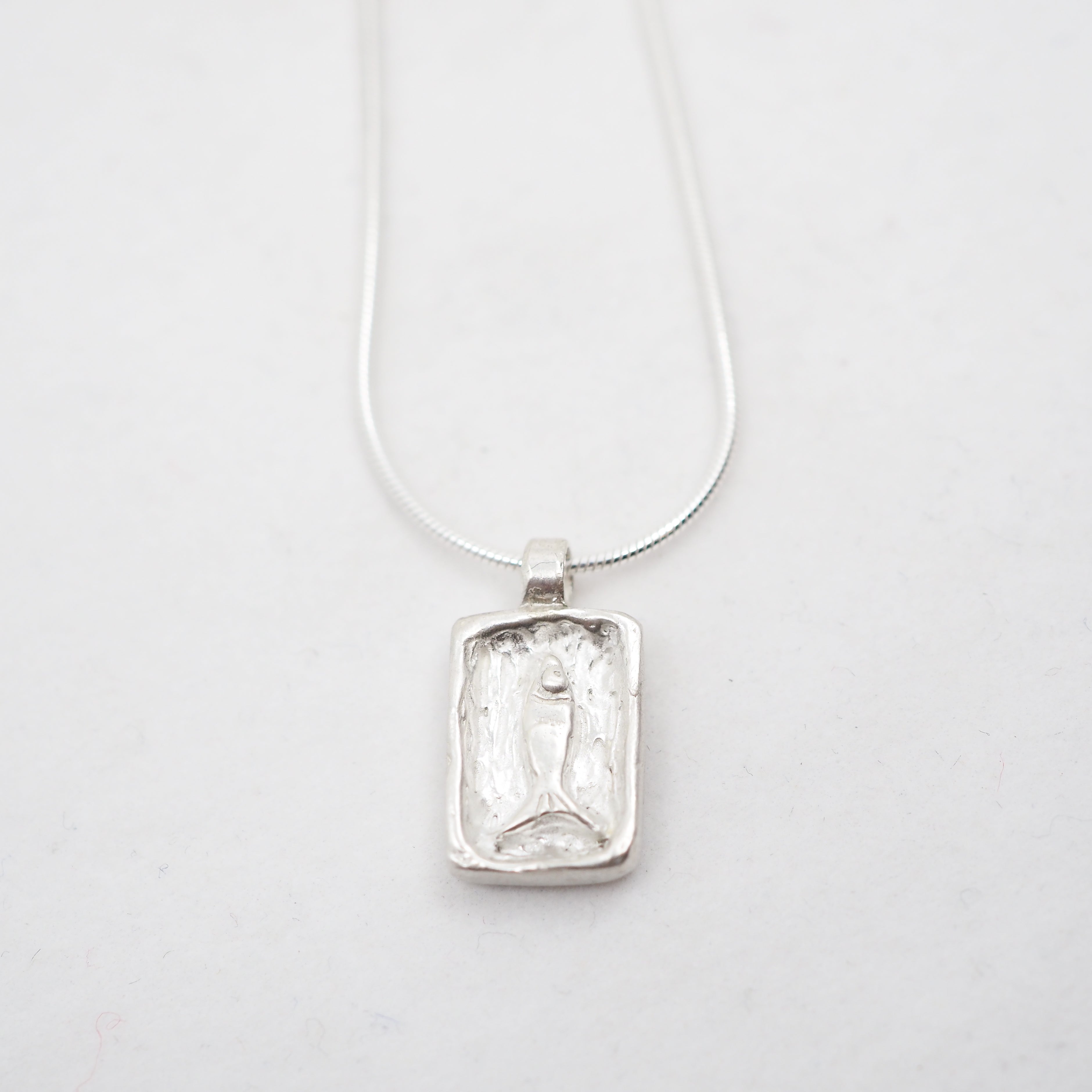 A silver rectangular pendant necklace featuring a textured, carved fish on a delicate silver chain, photographed on a clean white background.