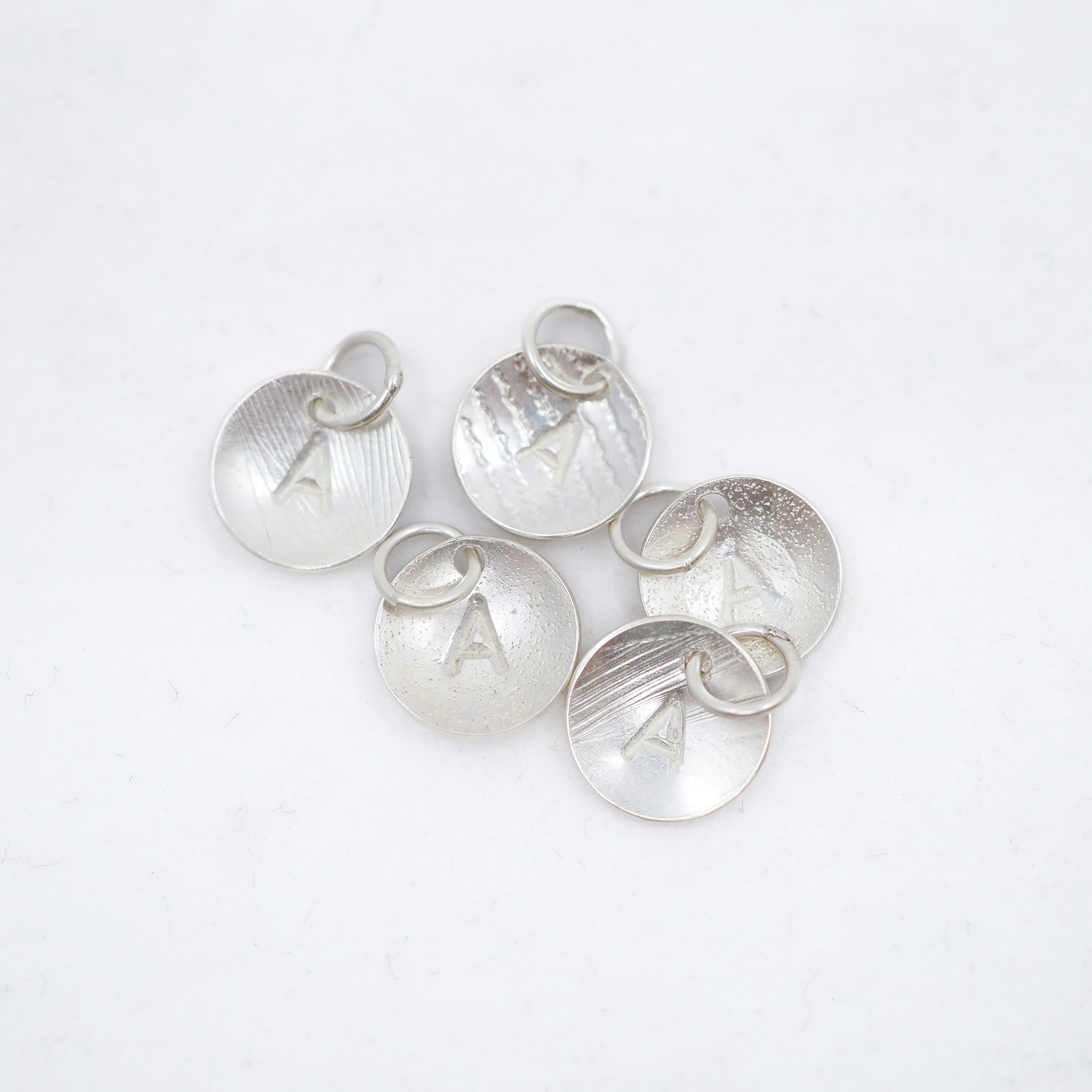 Five silver disc charms, each engraved with the letter A, arranged on a plain white background.