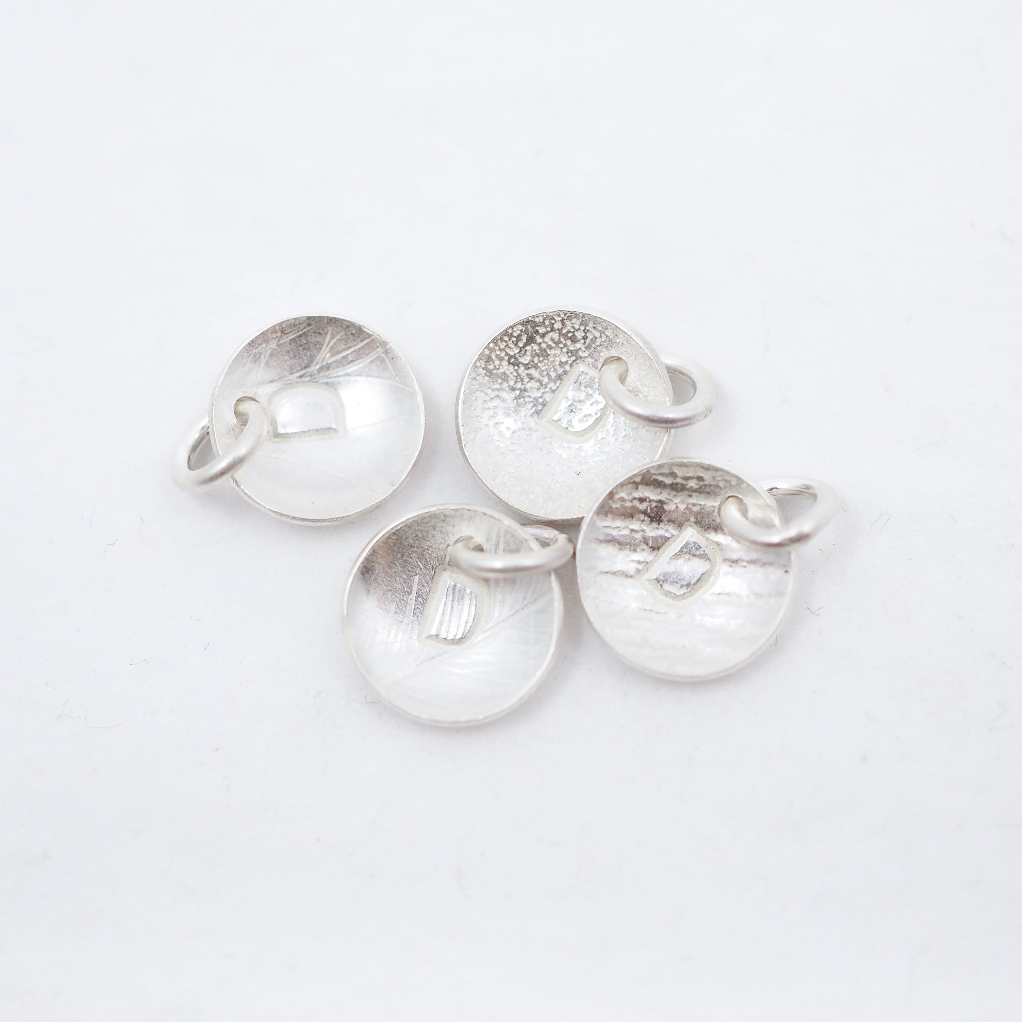 Four round silver disc charms with letter D textured finishes and small attached loops, arranged on a plain white background.