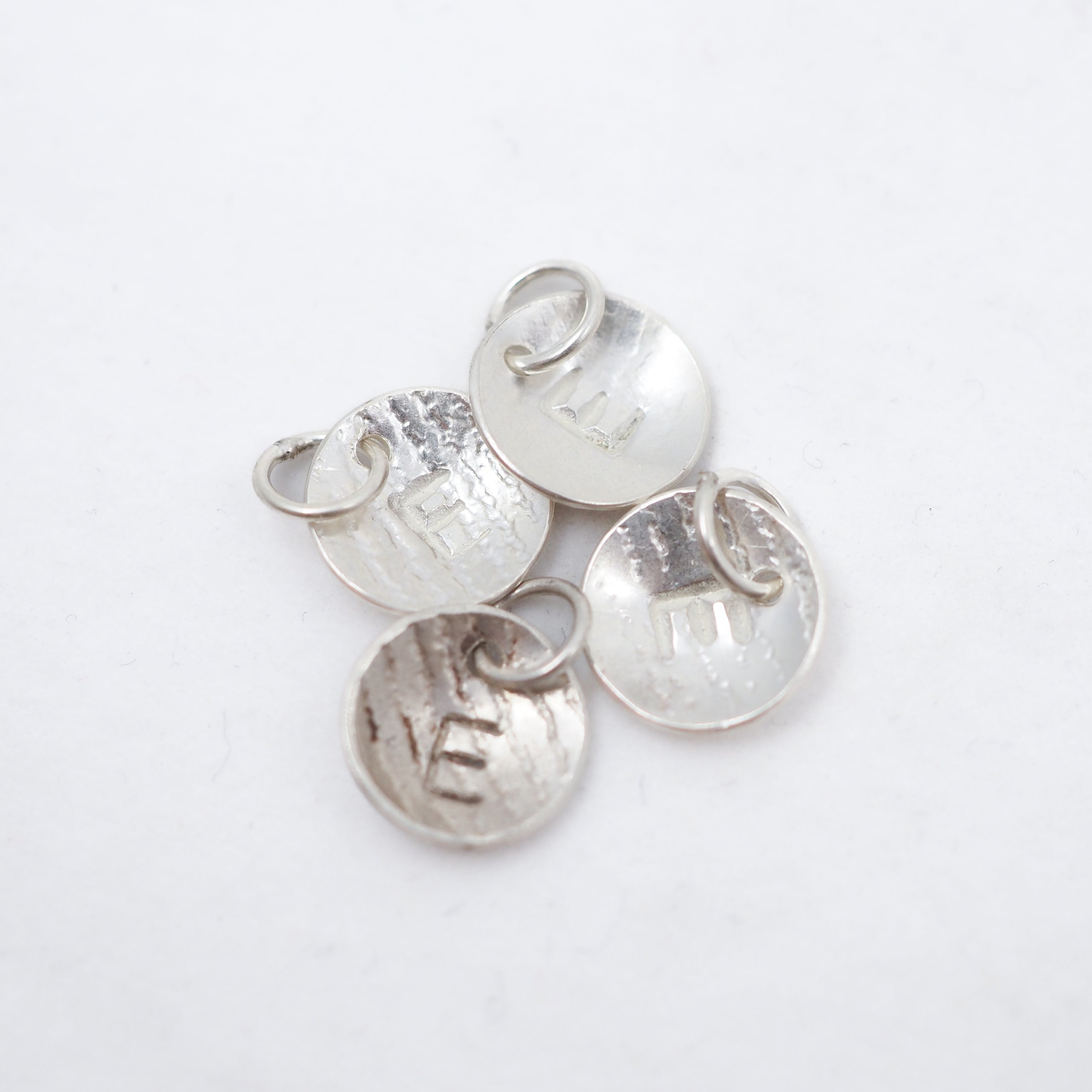 Four small round silver disc charms with attached jump rings, each stamped with the letter E, arranged on a plain white background.