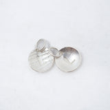 Two small round silver disc charms with textured and brushed finishes, one featuring a raised chevron and the other a heart cutout, shown on a white background.