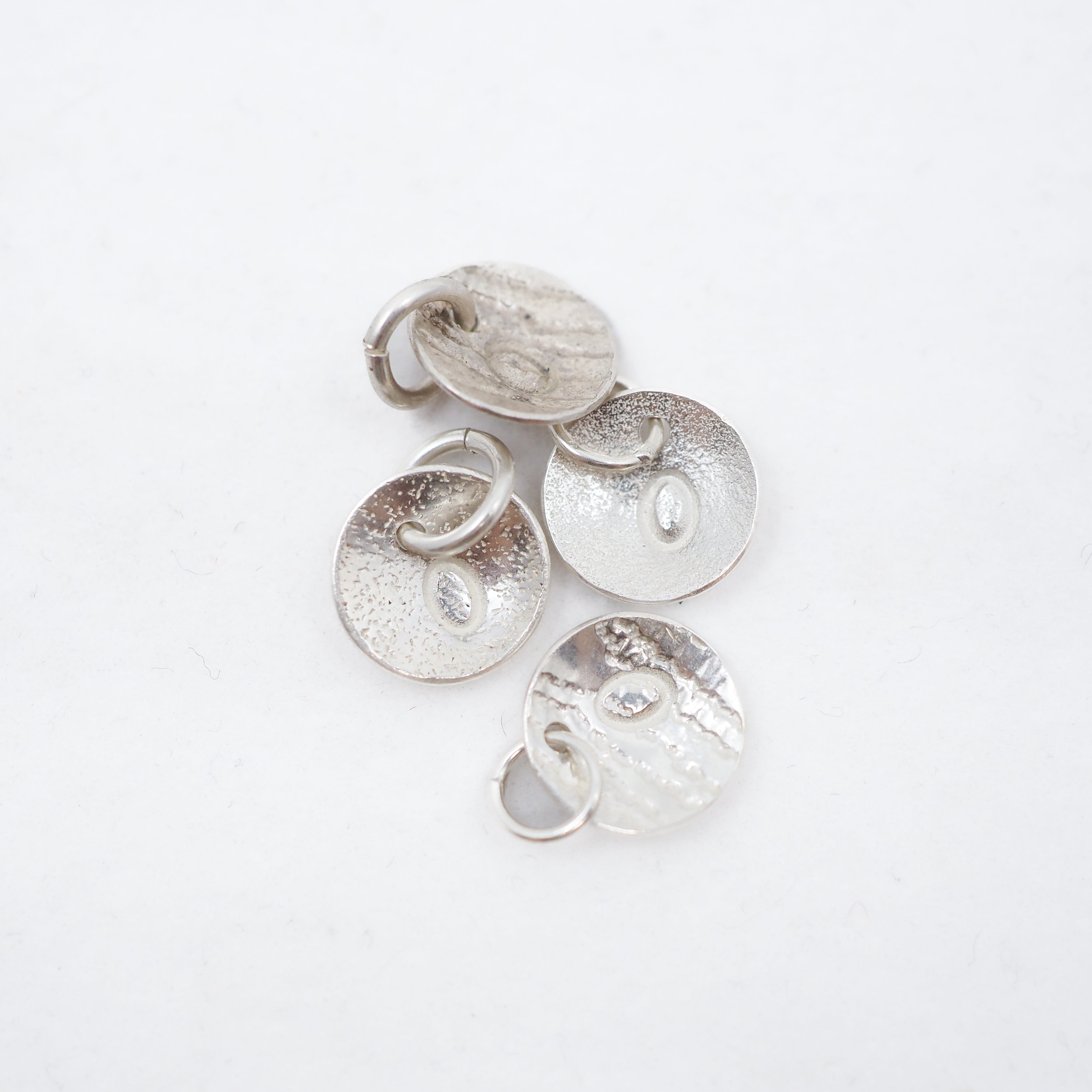 Four textured silver disc charms with small jump rings, arranged on a plain white background.