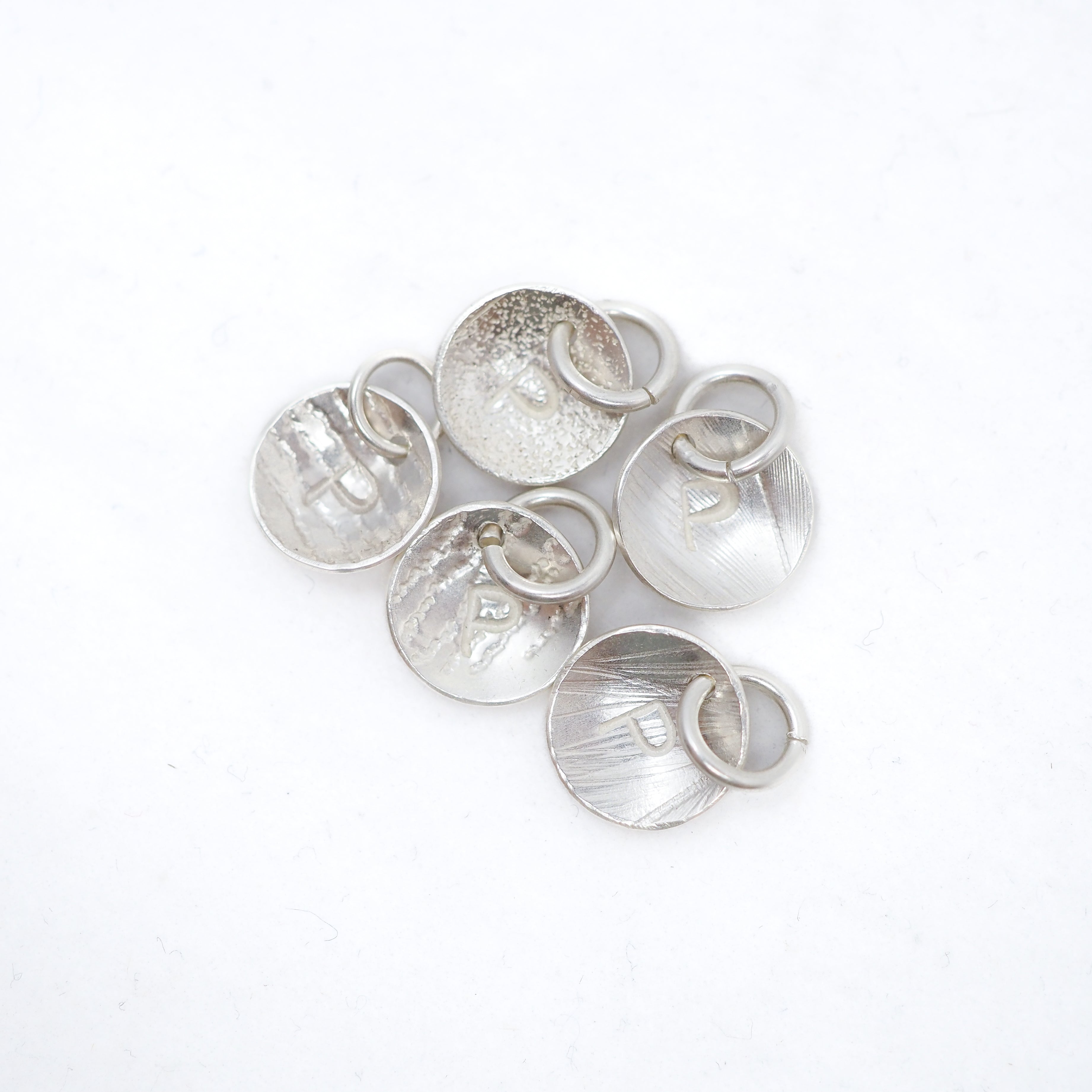 Five textured silver disc charms with attached small silver rings, arranged on a plain white background.