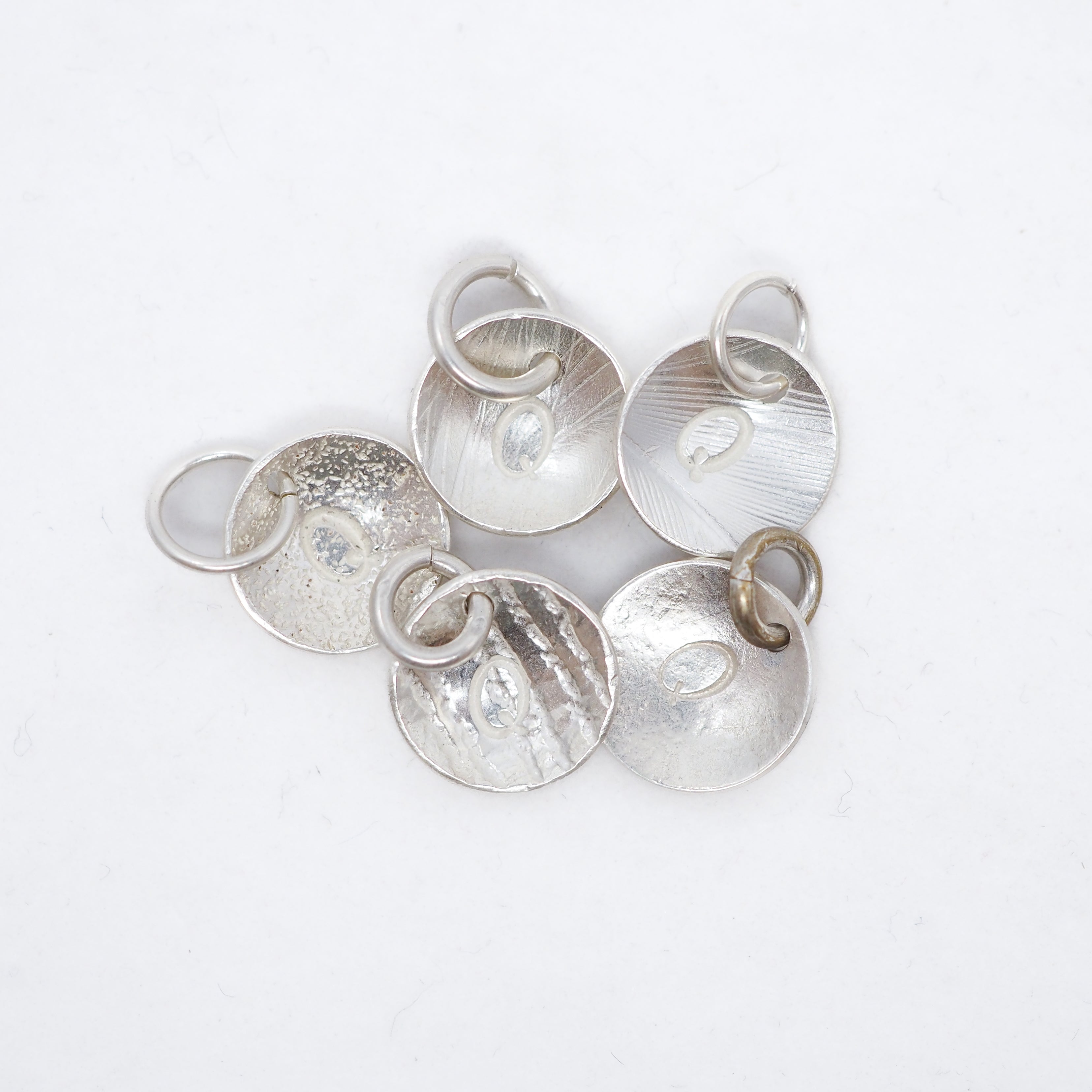 Five textured silver disc charms with attached jump rings, arranged on a plain white background.