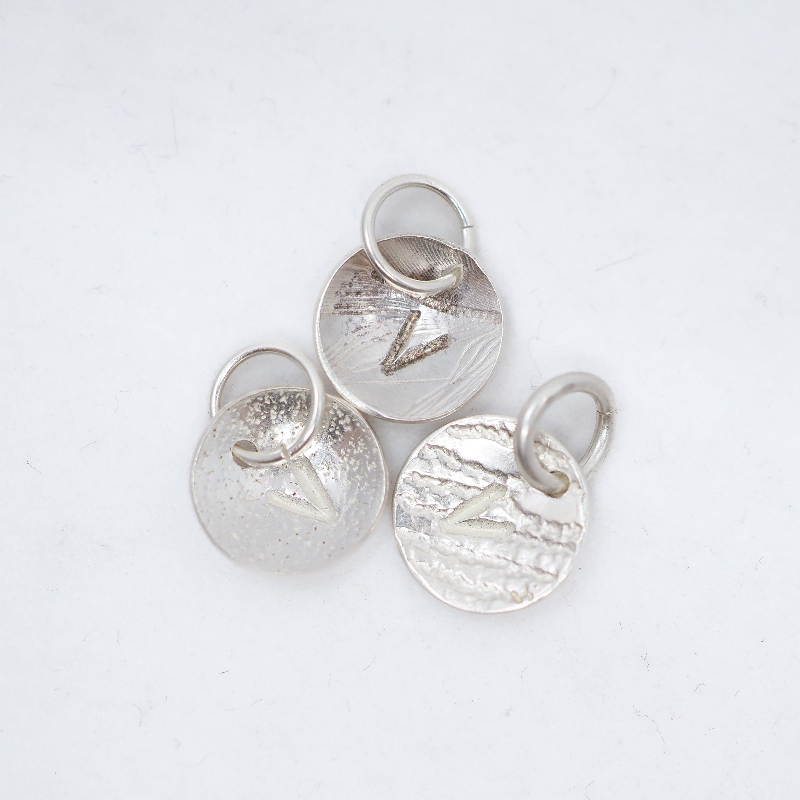 Three textured silver oval charms with jump rings, each featuring a different engraved letter V, arranged on a white background.
