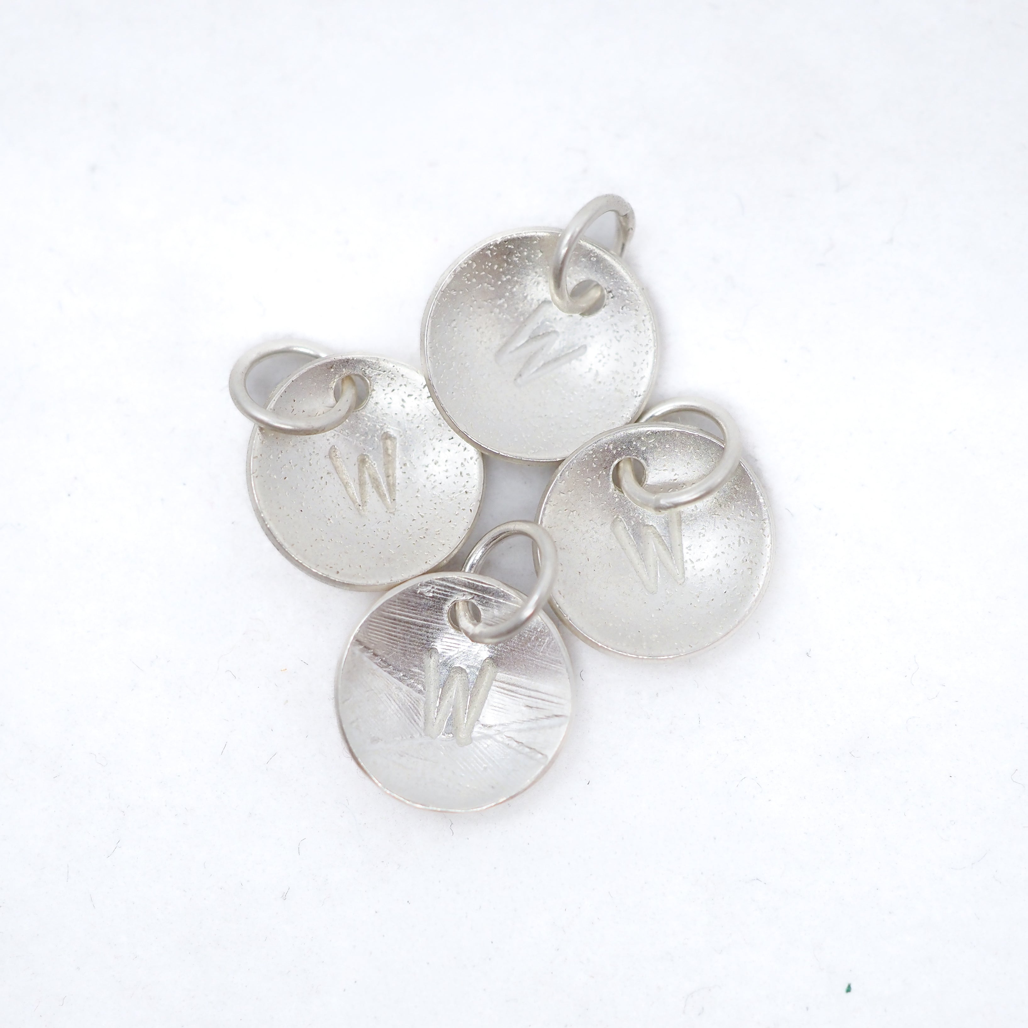 Four textured silver disc charms, each engraved with a letter 'W' and featuring a small jump ring, arranged on a plain white background.