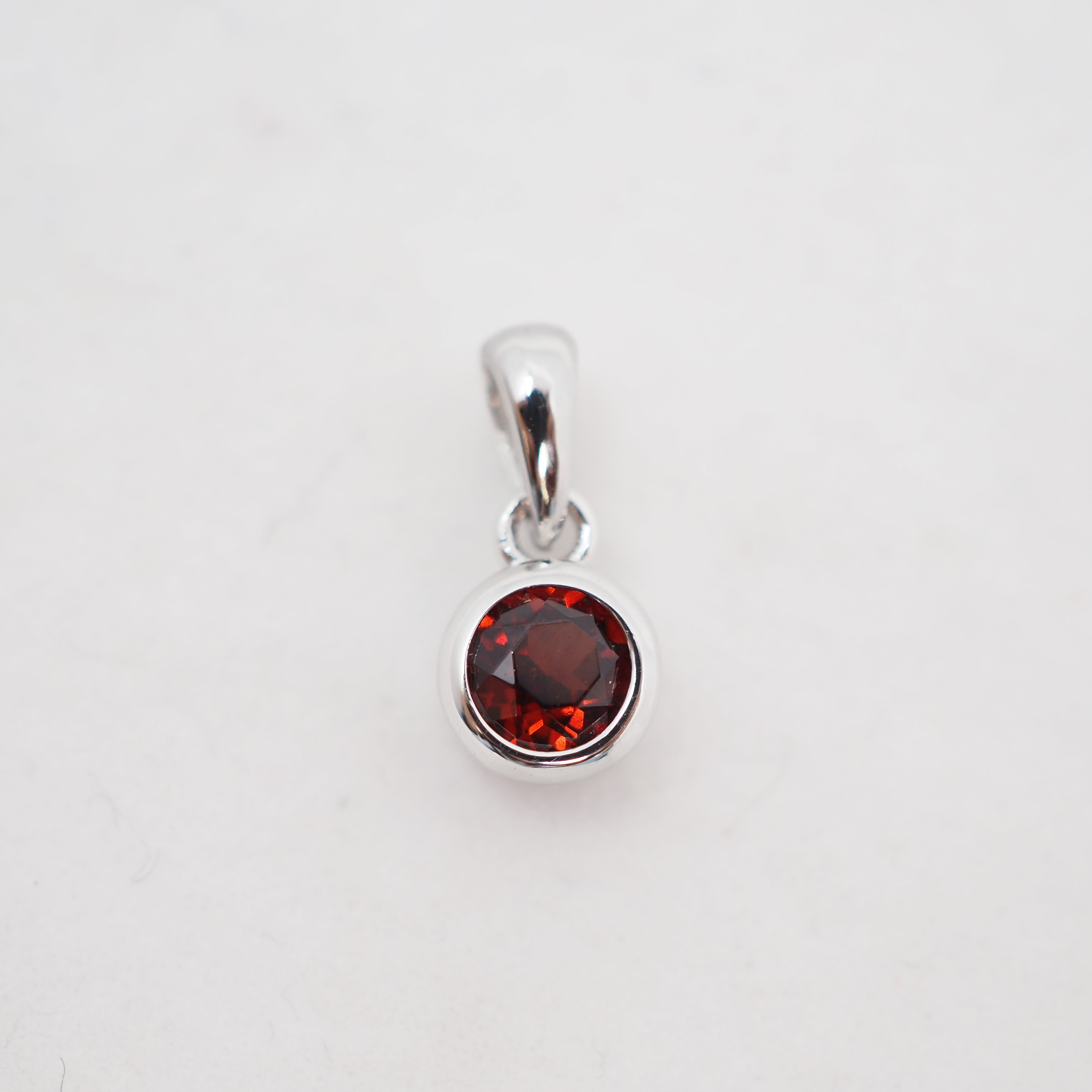 A sterling silver pendant charm featuring a round faceted dark red gemstone in a bezel setting, shown on a plain white background.