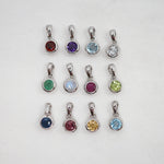 Twelve silver bezel-set gemstone pendants arranged in a 3-by-4 grid on a white background, featuring various colored stones including red, purple, light blue, clear, green, light blue, magenta, lime green, dark blue, yellow, magenta, and light blue.