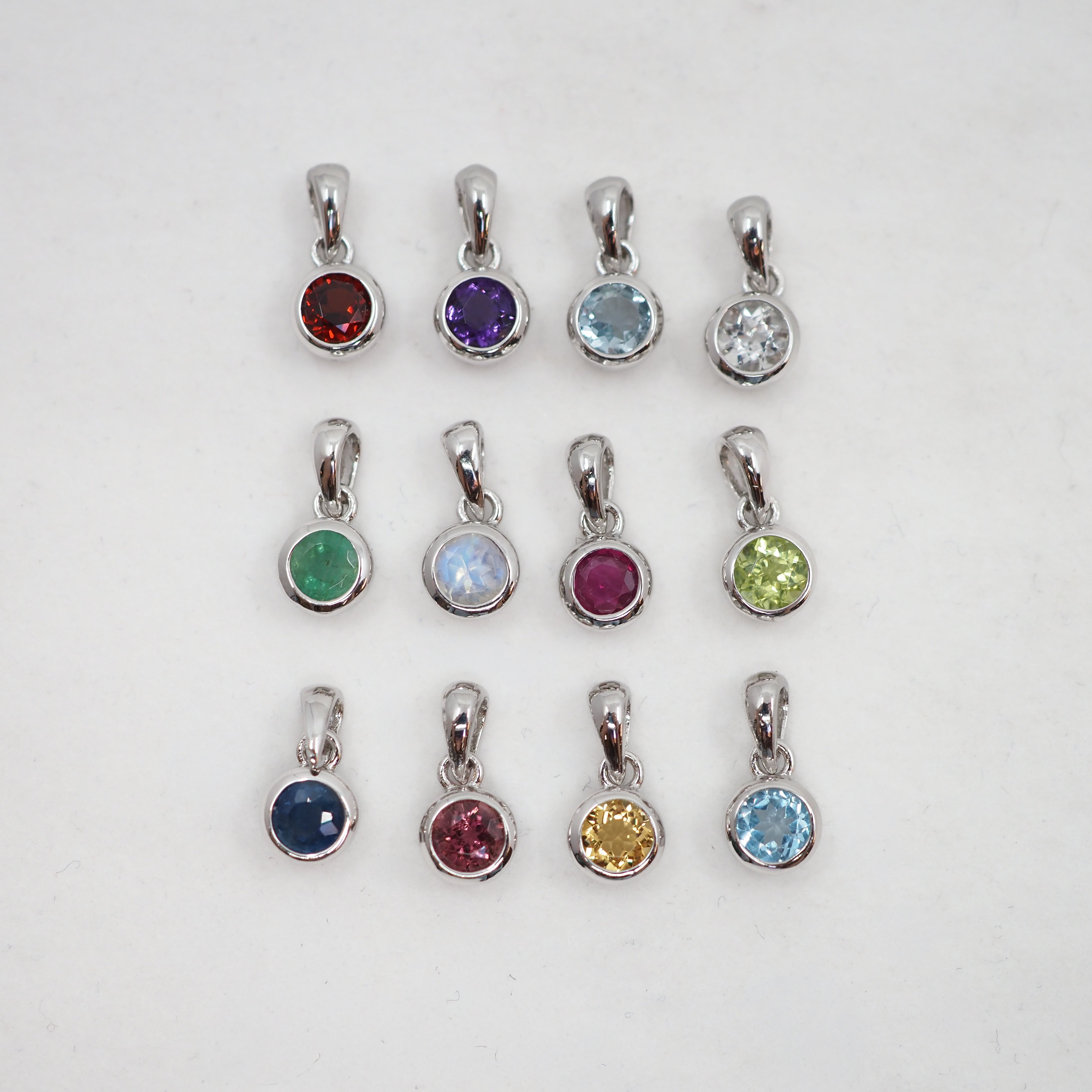 Twelve silver bezel-set gemstone pendants arranged in a 3-by-4 grid on a white background, featuring various colored stones including red, purple, light blue, clear, green, light blue, magenta, lime green, dark blue, yellow, magenta, and light blue.
