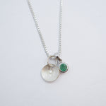 A silver chain necklace with two charms: a textured silver disc pendant and a small round green gemstone charm, displayed on a clean white background.
