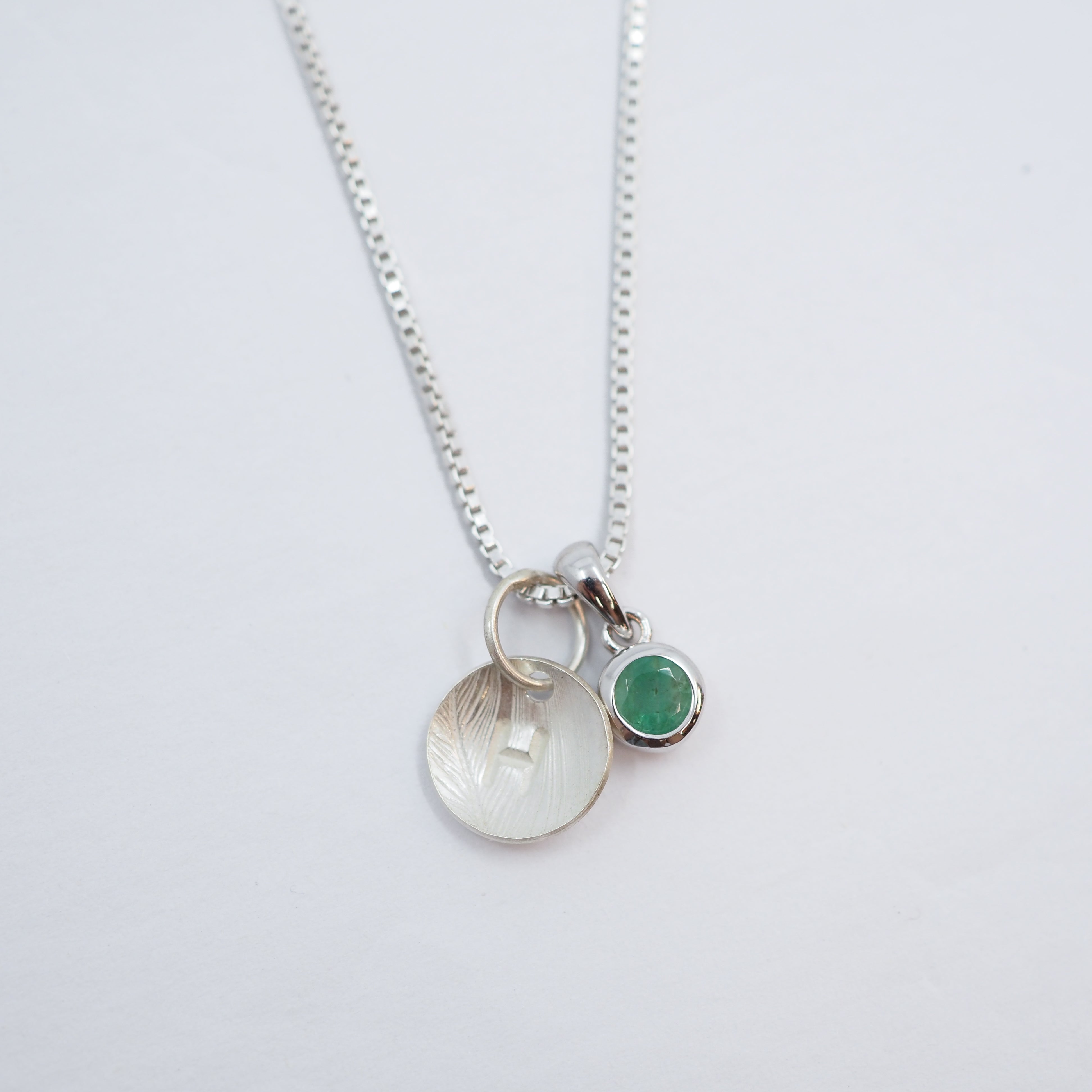 A silver chain necklace with two charms: a textured silver disc pendant and a small round green gemstone charm, displayed on a clean white background.