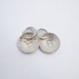 Two textured silver oval charms with a raised letter 'B' and jump rings, displayed on a clean white background.
