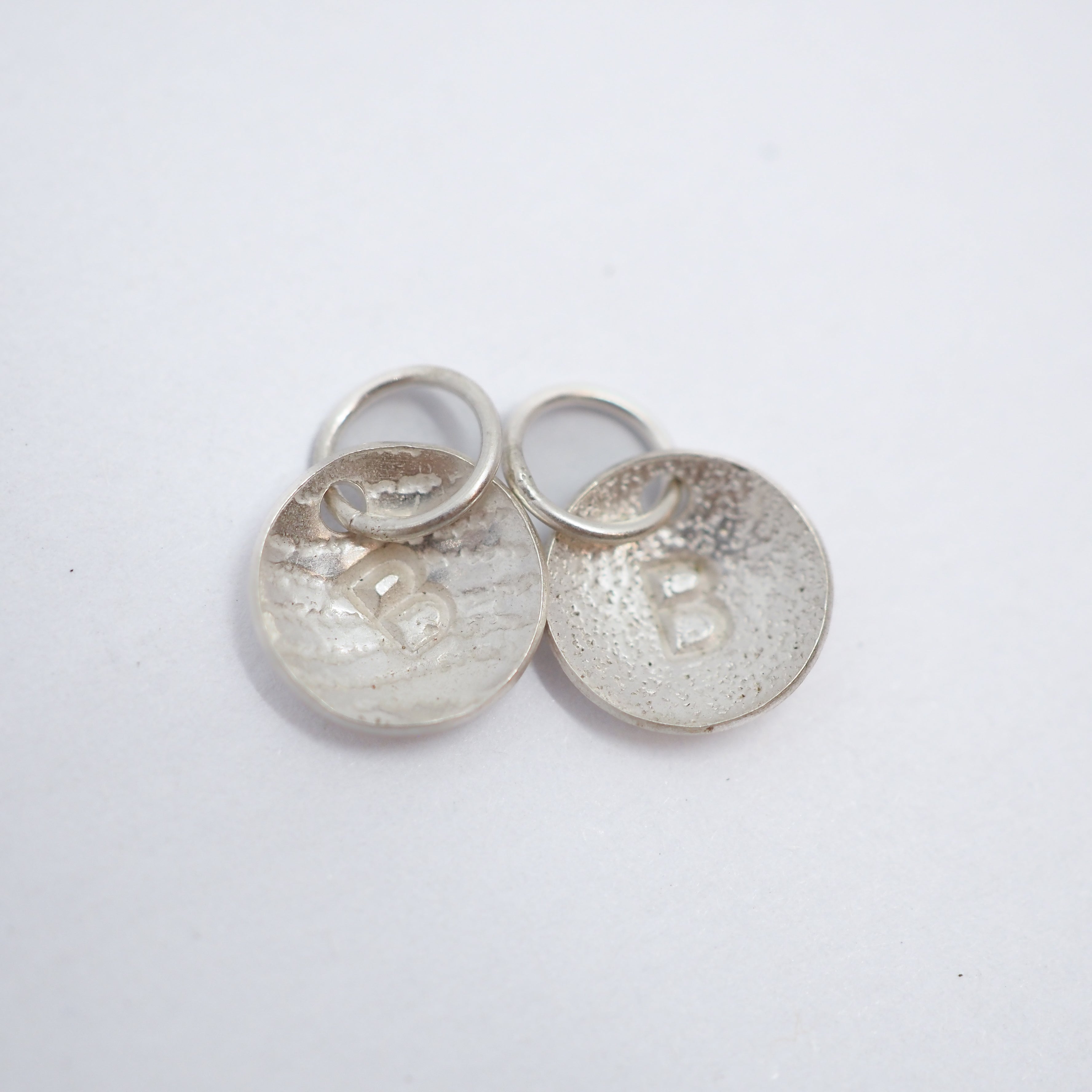 Two textured silver oval charms with a raised letter 'B' and jump rings, displayed on a clean white background.