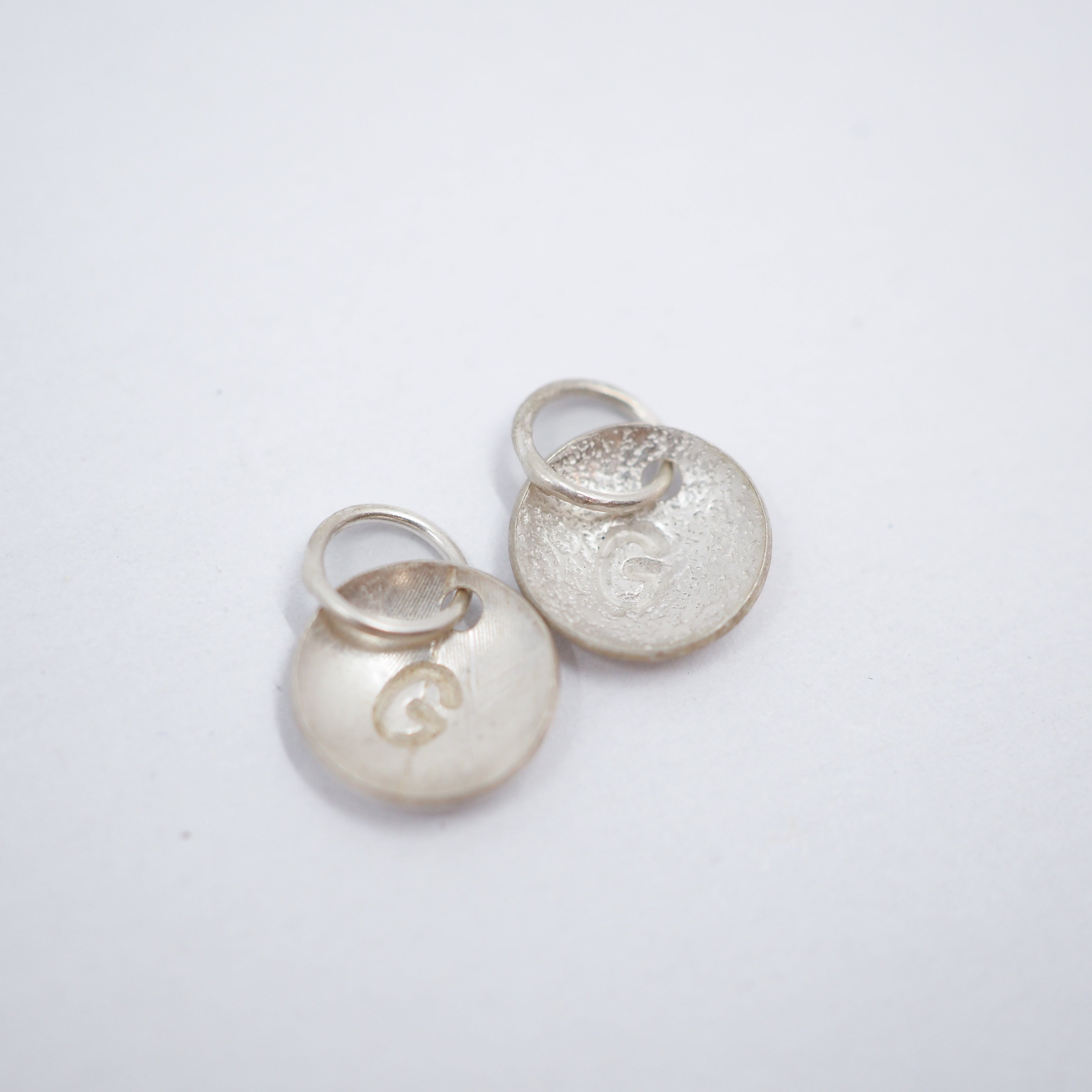 A small silver circular disc charm with a textured, hammered surface and a small attached metal loop, shown on a plain white background.