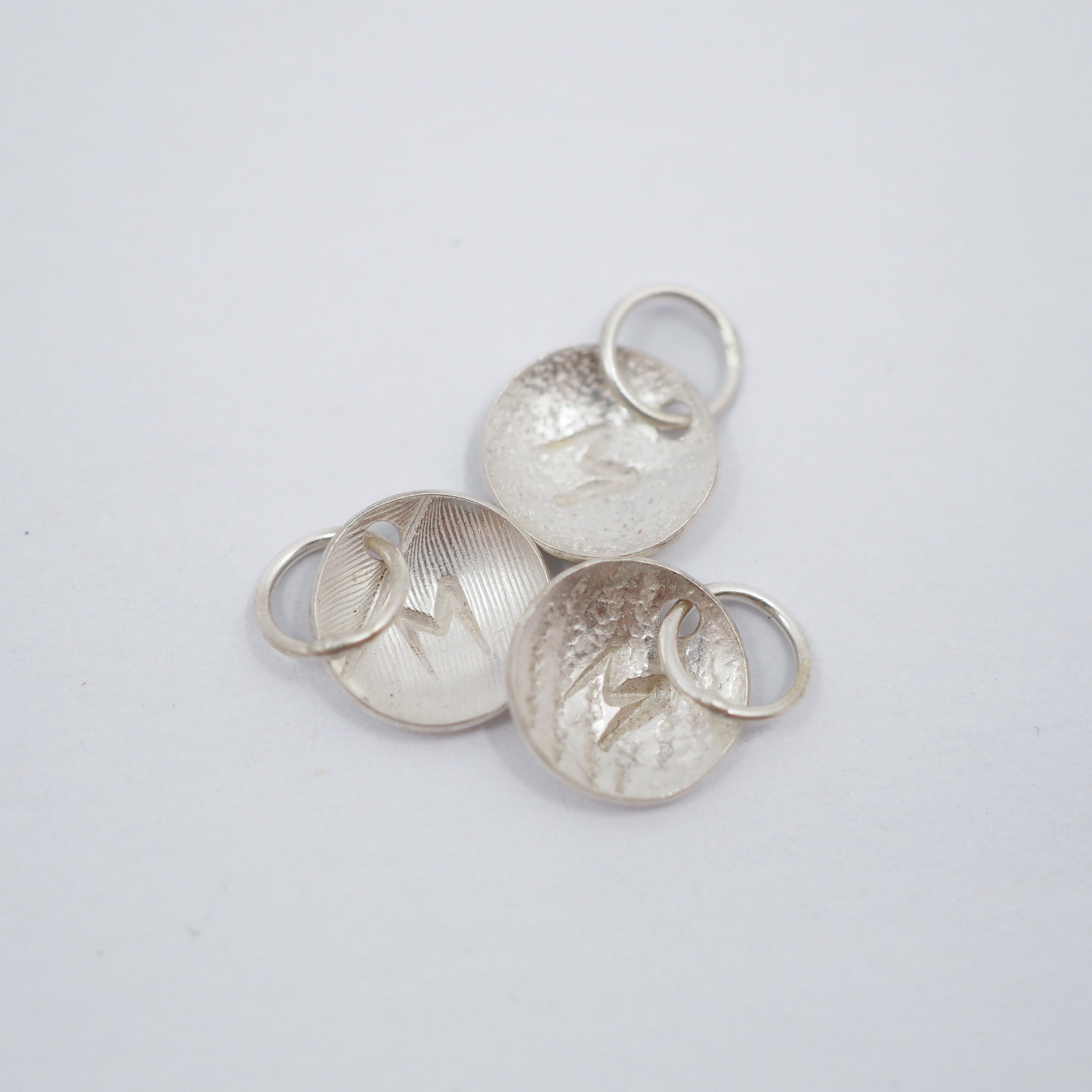 Three textured silver oval charms with lightning bolt details and attached split rings, arranged on a plain light gray background.