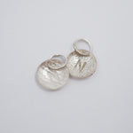 Two textured silver disc charms with engraved letter 'N' and jump rings, displayed on a light gray background.
