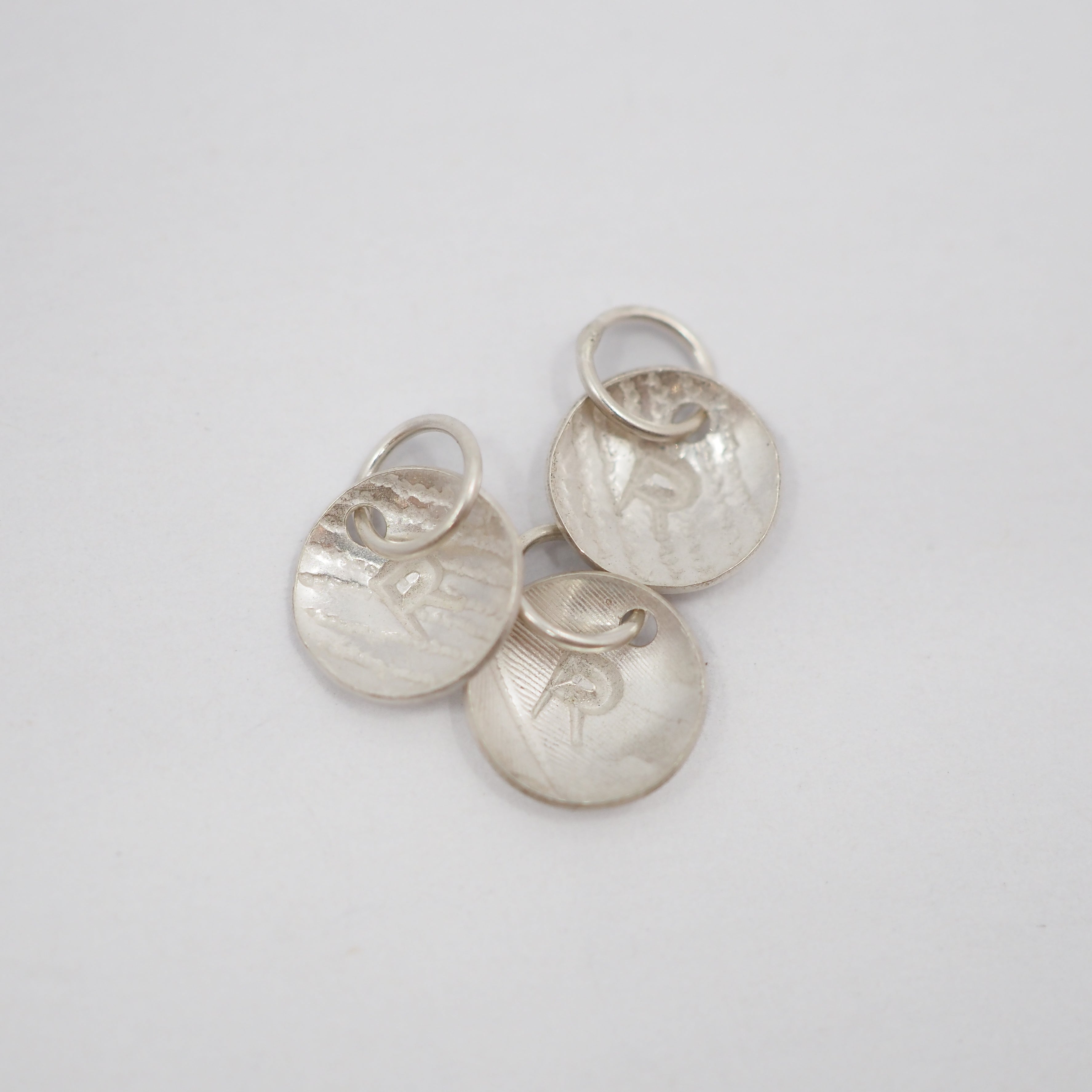 Three small hammered silver oval charms with attached loops, arranged on a plain light gray background.