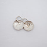 Two small silver oval charms with jump rings, one brushed with a cross and the other textured with a cross, on a light gray background.