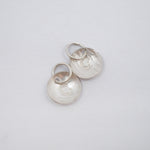 Two silver disc charms with a hammered texture and a small heart cutout, attached to silver jump rings, shown on a plain light gray background.
