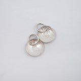 Two silver disc charms with a hammered texture and a small heart cutout, attached to silver jump rings, shown on a plain light gray background.