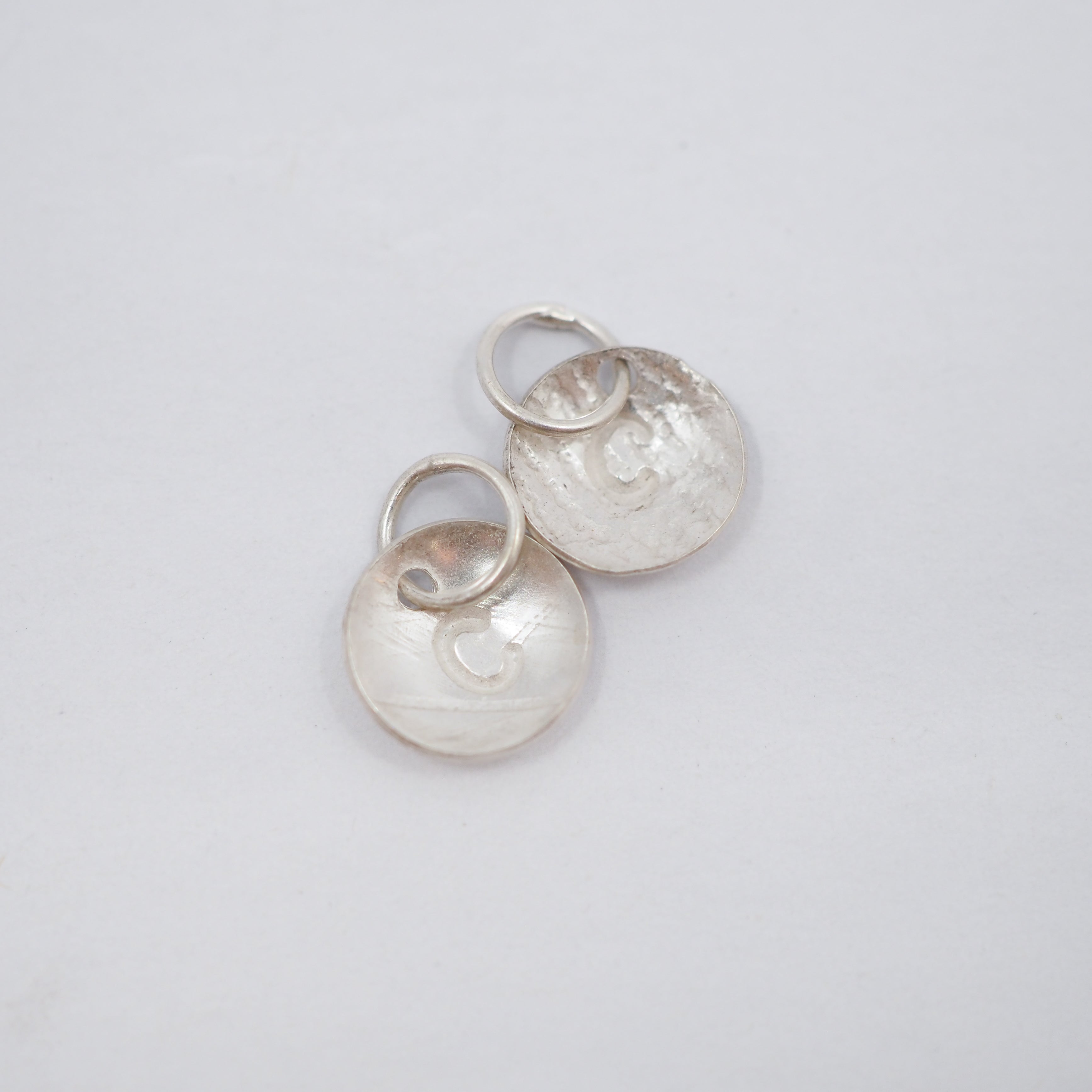 Two silver disc charms with a hammered texture and a small heart cutout, attached to silver jump rings, shown on a plain light gray background.