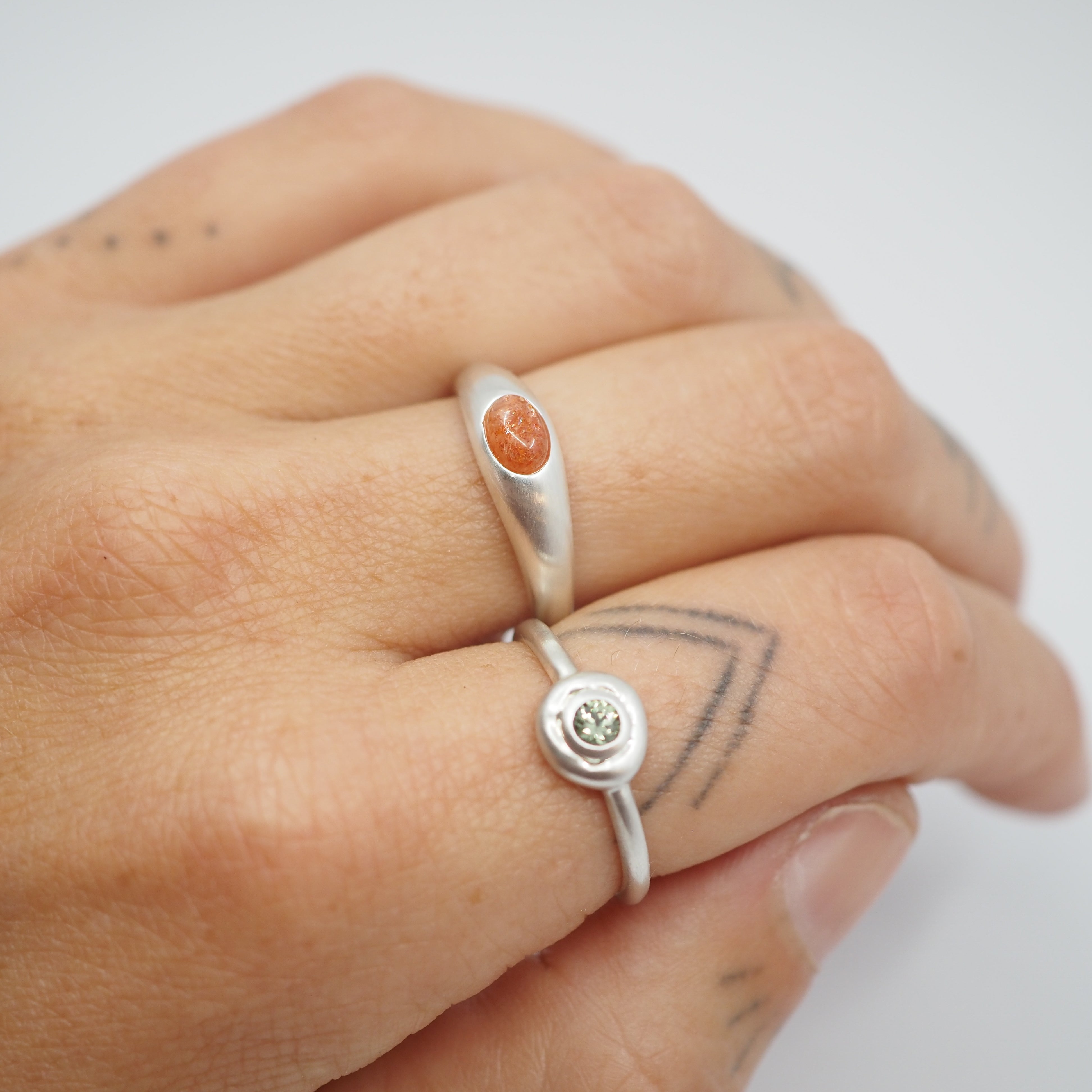 A close-up of a hand wearing two silver rings. The top ring has a smooth silver band with a small oval orange stone, and the bottom ring has a thin silver band with a round bezel-set light green gemstone.
