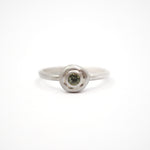 A silver ring with a textured, circular bezel setting holding a small round green gemstone, photographed on a seamless white background.