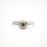 A silver ring with a textured, circular bezel setting holding a small round green gemstone, photographed on a seamless white background.