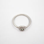 A minimalist silver ring with a small oval bezel containing a clear round gemstone, set against a plain white background.