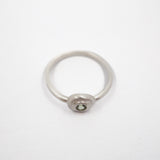 A minimalist silver ring with a small oval bezel containing a clear round gemstone, set against a plain white background.