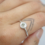 A close-up of a hand wearing a silver ring with a small round clear gemstone, showing the ring on the middle finger against a light background.